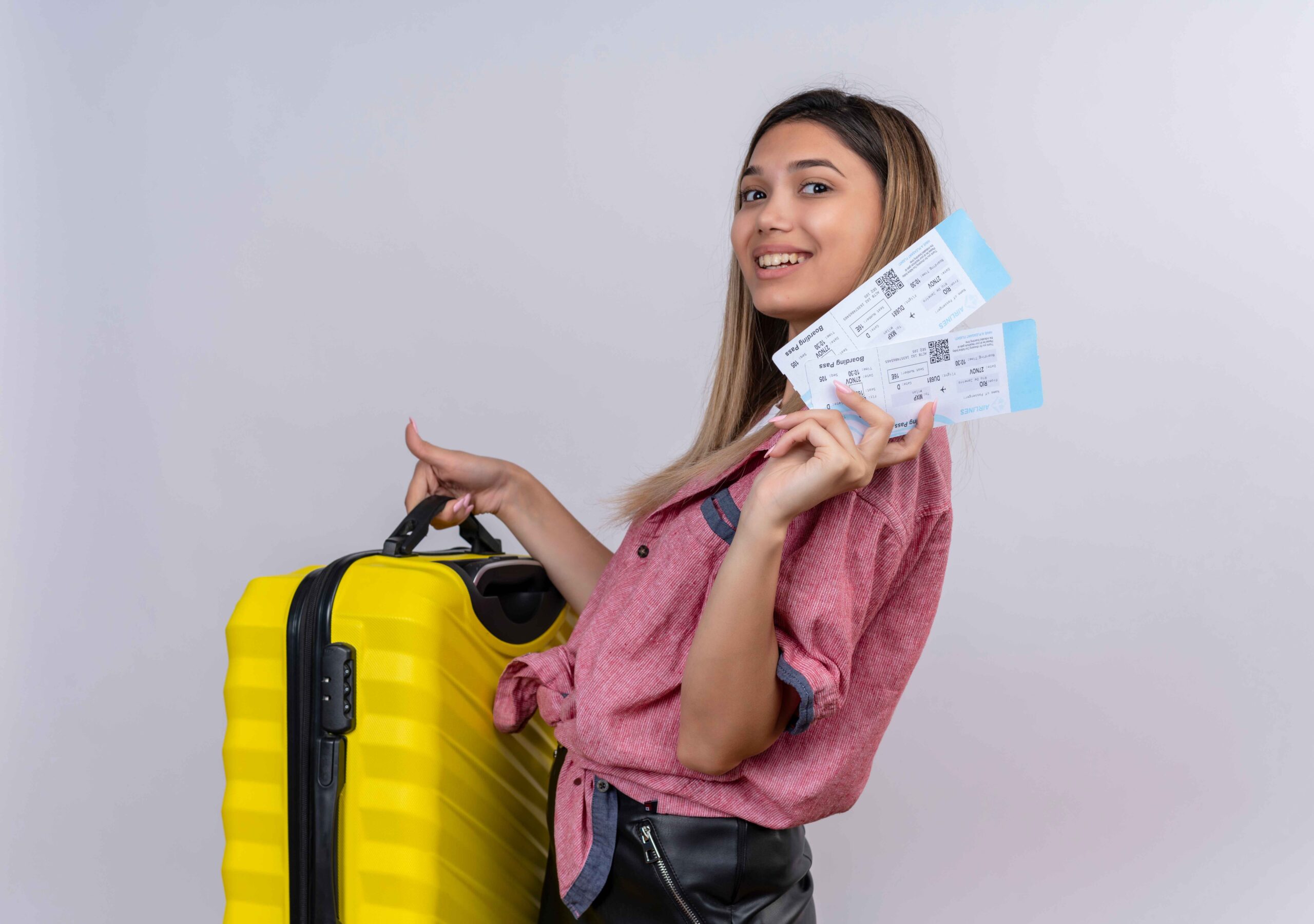 pleased young woman wearing red shirt holding yellow suitcase with plane tickets while looking white wall(1)